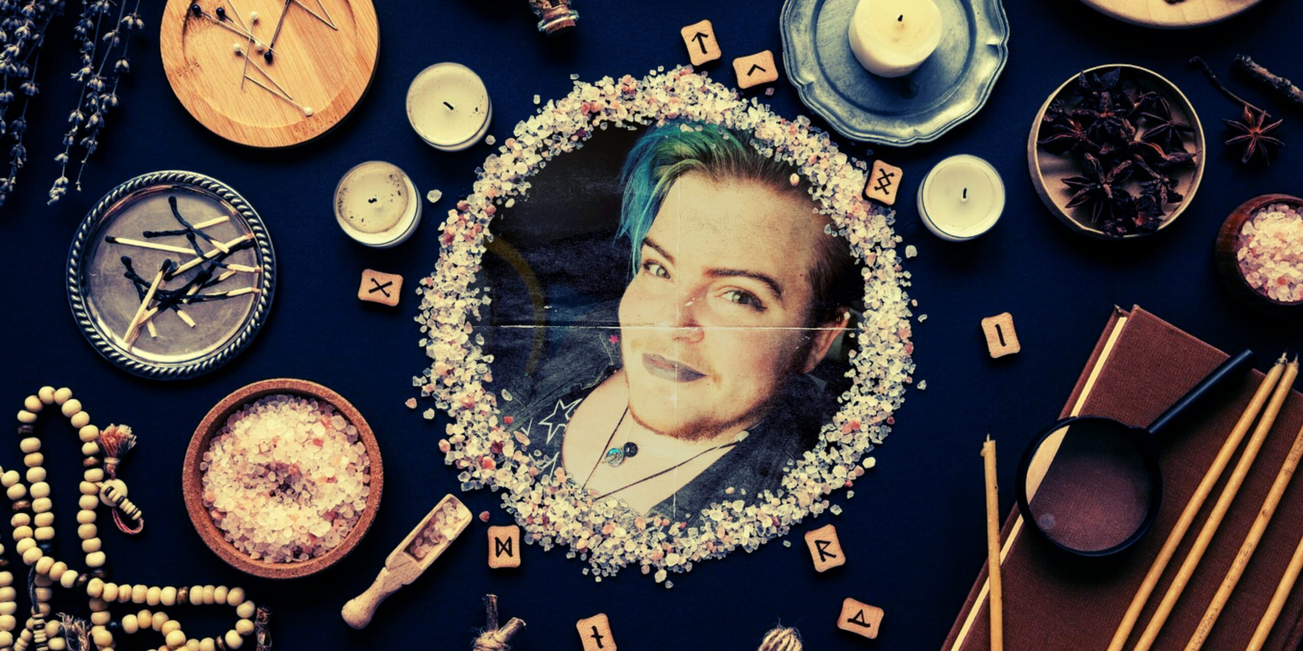 Photo of a table of esoterica, like candles, used matches, prayer beads, some varieties of herbs and fungi, some wooden runes, and a circle of salt in the center. Inside the circle of salt is a creased paper printout of Chiron's face. They are a white transmasc queer with a beard and blue hair, wearing taupe lipstick, a denim jacket, and a raven pendant.